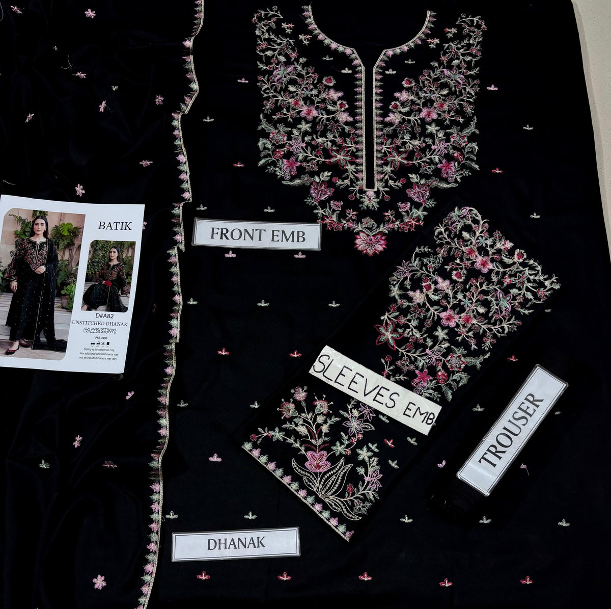 Black embroidered ethnic suit fabric set with floral embroidery, shown laid out with labels for front embroidery, sleeves, trouser, and dhanak. A small printed card is visible on the left.