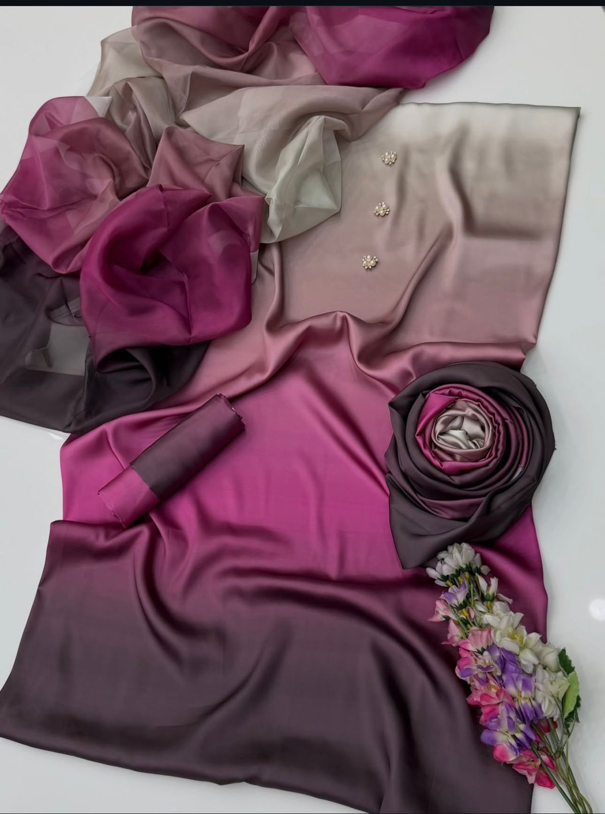 A draped satin scarf or shawl with a smooth ombré color transition from deep magenta and plum purple to taupe and dark charcoal gray, styled with small gold floral embellishments and a small bouquet of flowers on a white background.