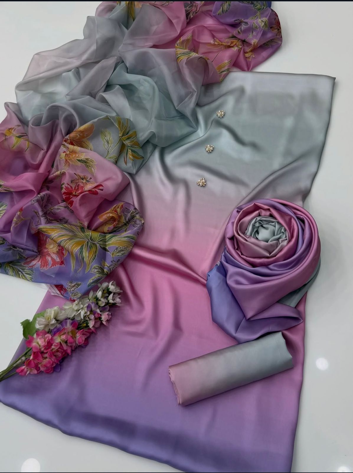 A folded and draped satin fabric set with a smooth gradient transitioning from light blue-gray to pink and lavender, accented with a matching sheer floral scarf and small decorative flower petals on a white background.