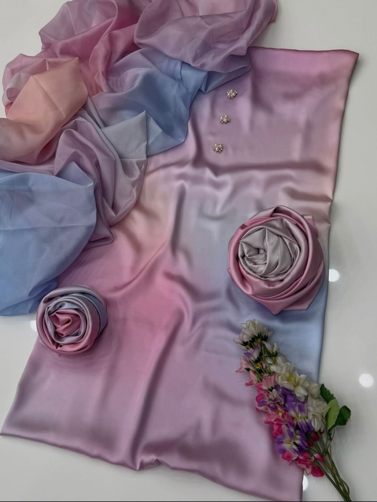 A draped satin fabric with a pink and light blue gradient, styled with two rolled fabric roses, small gold floral embellishments, and a small bouquet of flowers on a white surface.