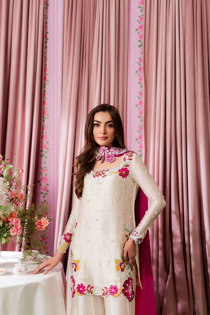 Woman in a white embroidered outfit standing in front of pink curtains with floral decorations.