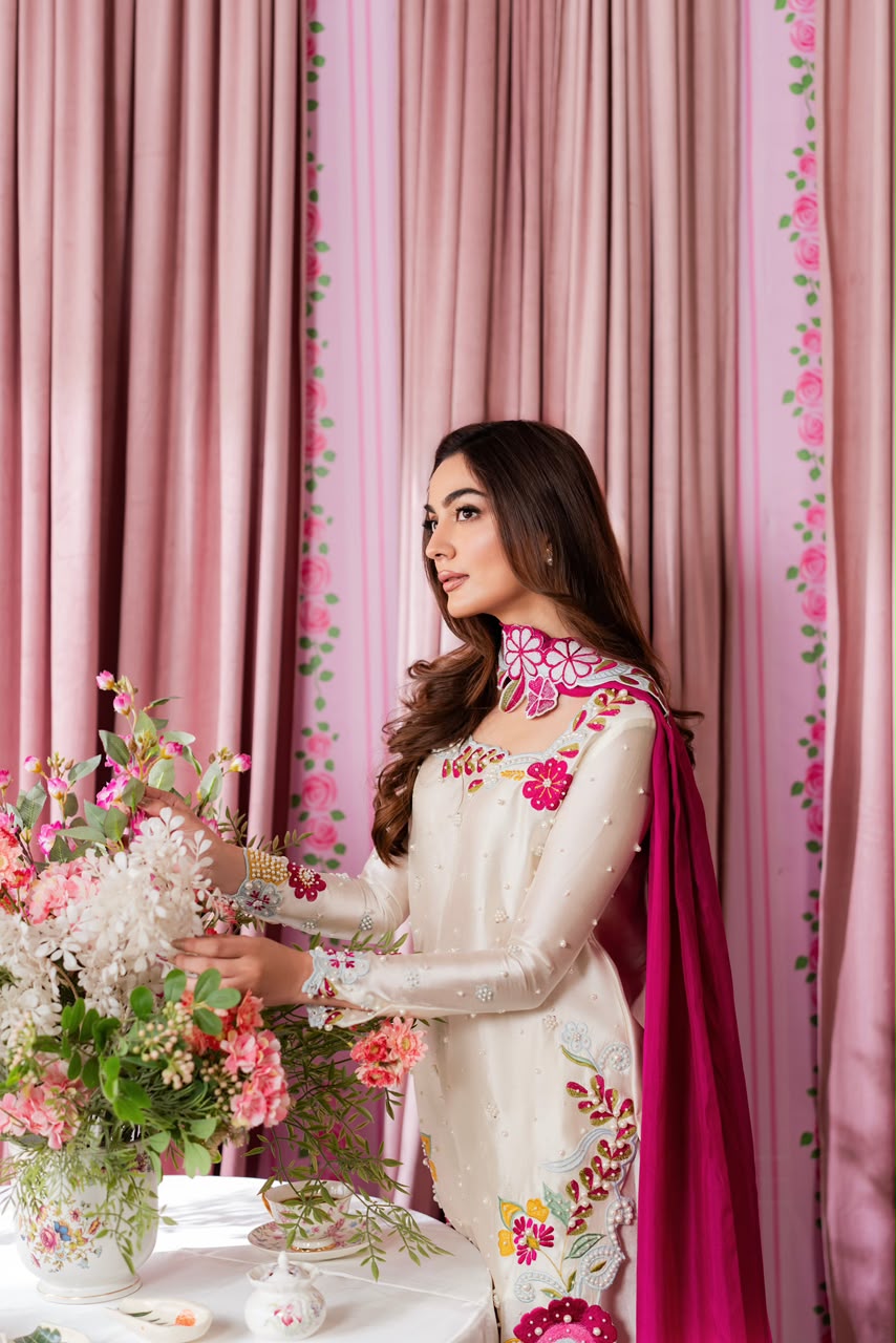 A woman wearing a cream-colored embroidered traditional outfit with a bright pink dupatta stands beside a table with a large floral arrangement, set against pink patterned curtains.
