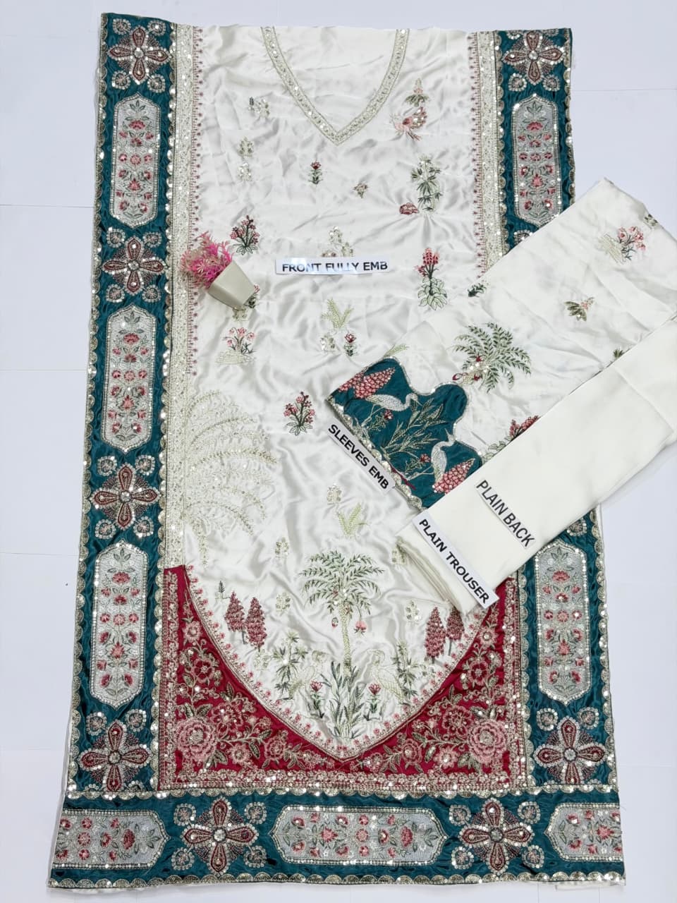 An embroidered white salwar suit set laid flat on a white background, featuring a kurta with intricate floral embroidery, a matching flat skirt, and a pair of trousers. The fabric has detailed teal borders and red floral patterns.
