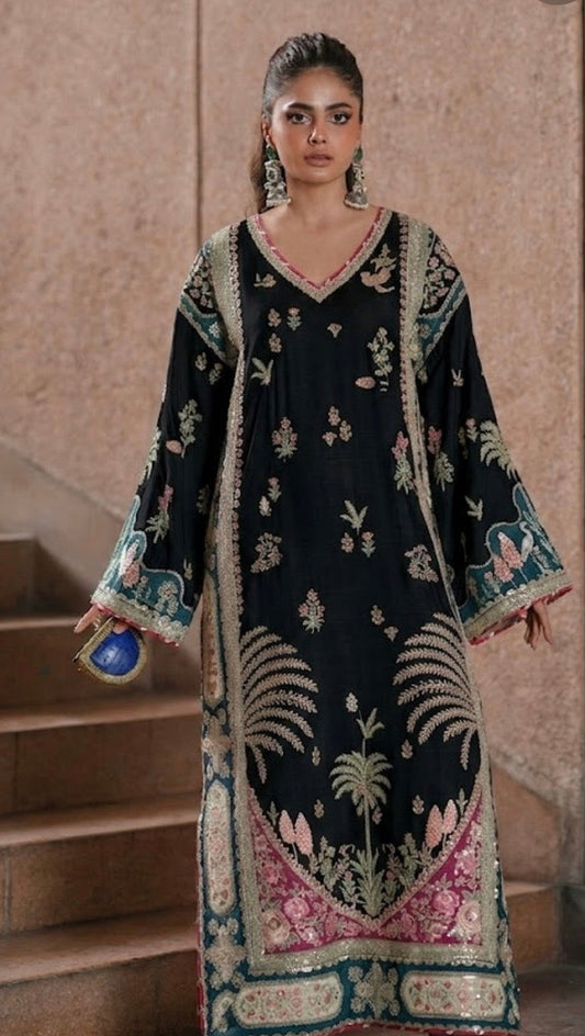 A woman models a black traditional long kurta with intricate multicolor embroidery, standing on stone steps against a textured beige wall.