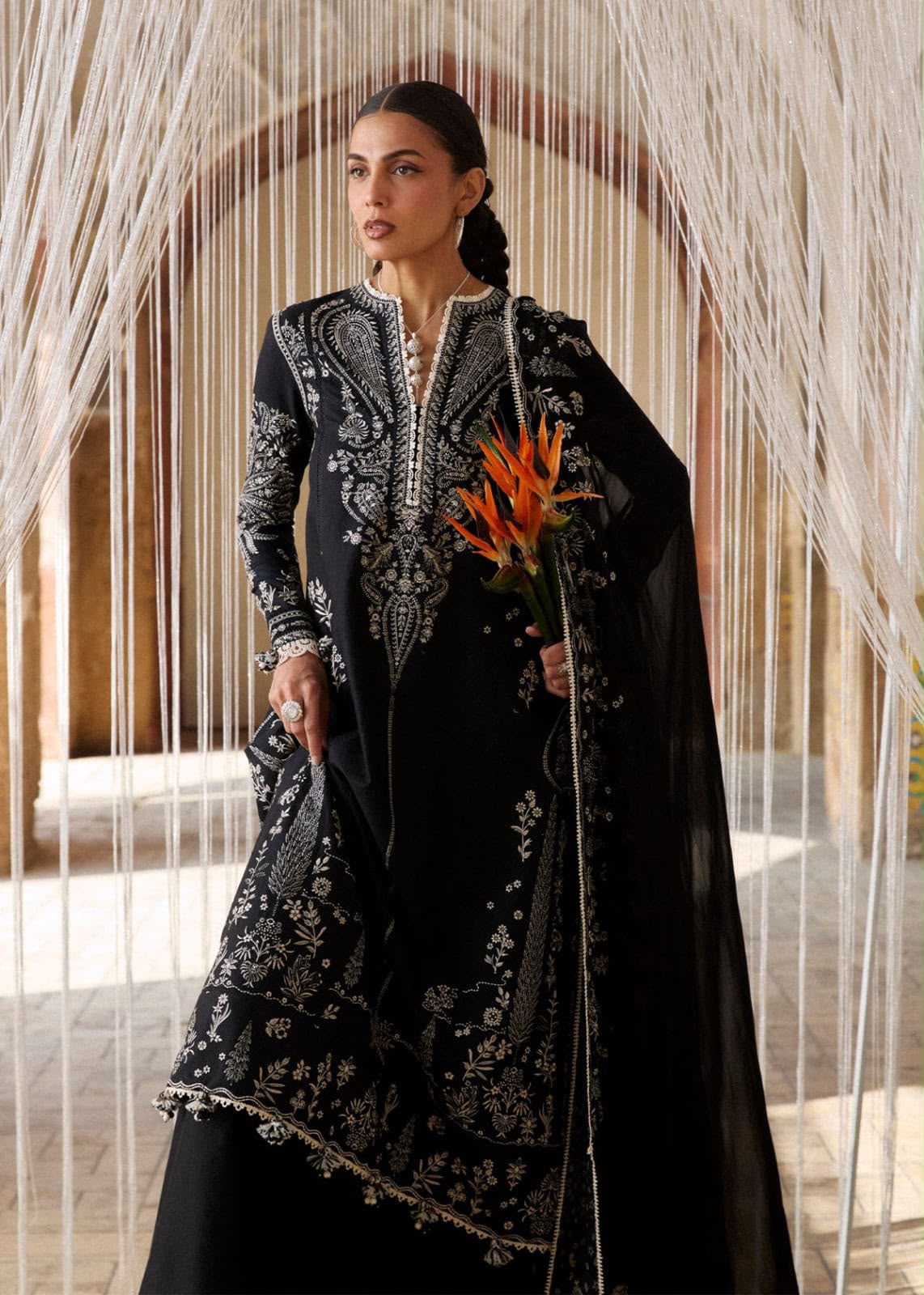 A woman models a black traditional outfit with intricate silver embroidery, featuring a long kurta, matching skirt, and a sheer black dupatta, while holding a vibrant orange flower against an arched background with hanging white strands.