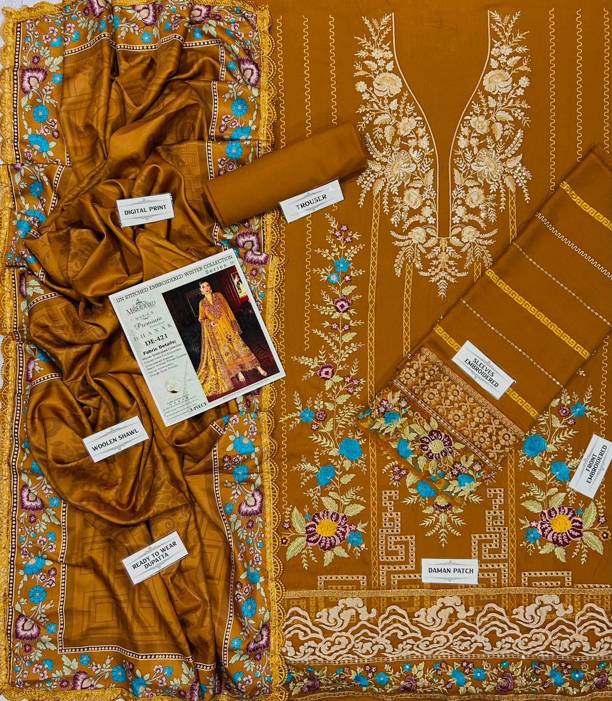 A folded and draped three-piece women's salwar suit set in a rich mustard-gold color, featuring intricate white, blue, and purple floral embroidery. The set includes a kurta, matching trousers, and a dupatta, with brand labels and a style card visible.