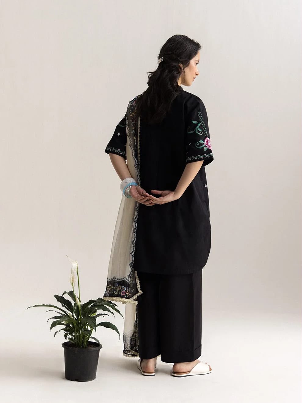 A woman stands in profile wearing a black traditional outfit with embroidered sleeves and a light beige patterned dupatta, photographed against a plain light beige studio background with a small potted plant at the lower left.