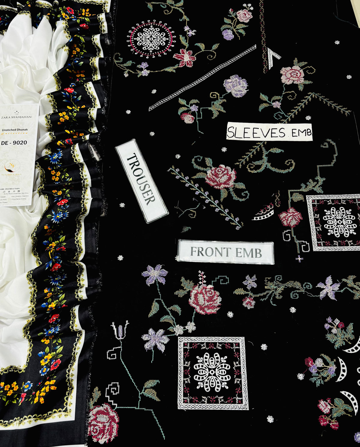 A close-up of a black embroidered garment or fabric sample laid flat, showing floral embroidery in pink, purple, green, and white, plus labels reading TROUSER, SLEEVES EMB, and FRONT EMB. A white ruffled trim with multicolor floral embroidery is visible along the left edge.