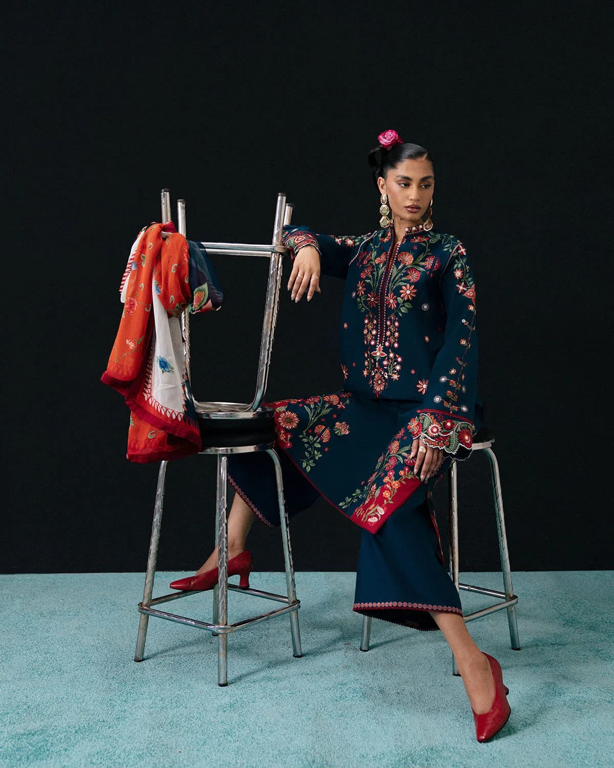 Woman in a floral embroidered outfit sitting on a stool with a black background