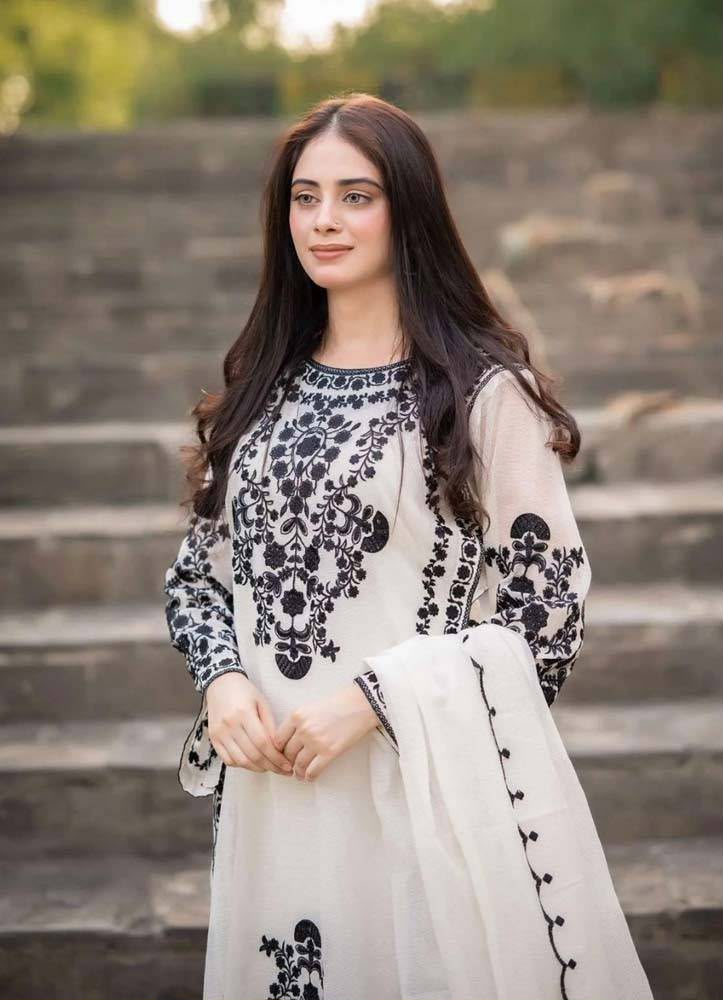 A woman stands on outdoor stone steps wearing a white traditional outfit with black floral embroidery and a matching white dupatta.