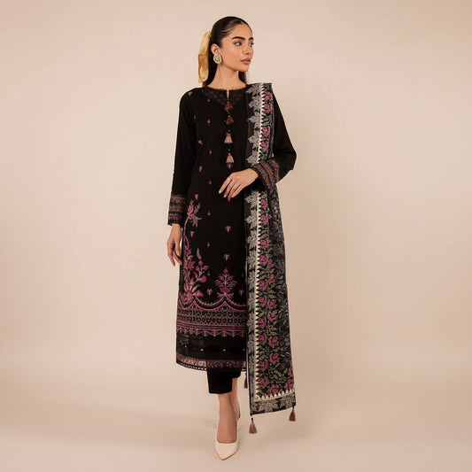 A woman models a black three-piece ethnic suit with intricate pink and gold embroidery, featuring a long kurta, matching trousers, and a patterned dupatta, set against a warm beige interior background.