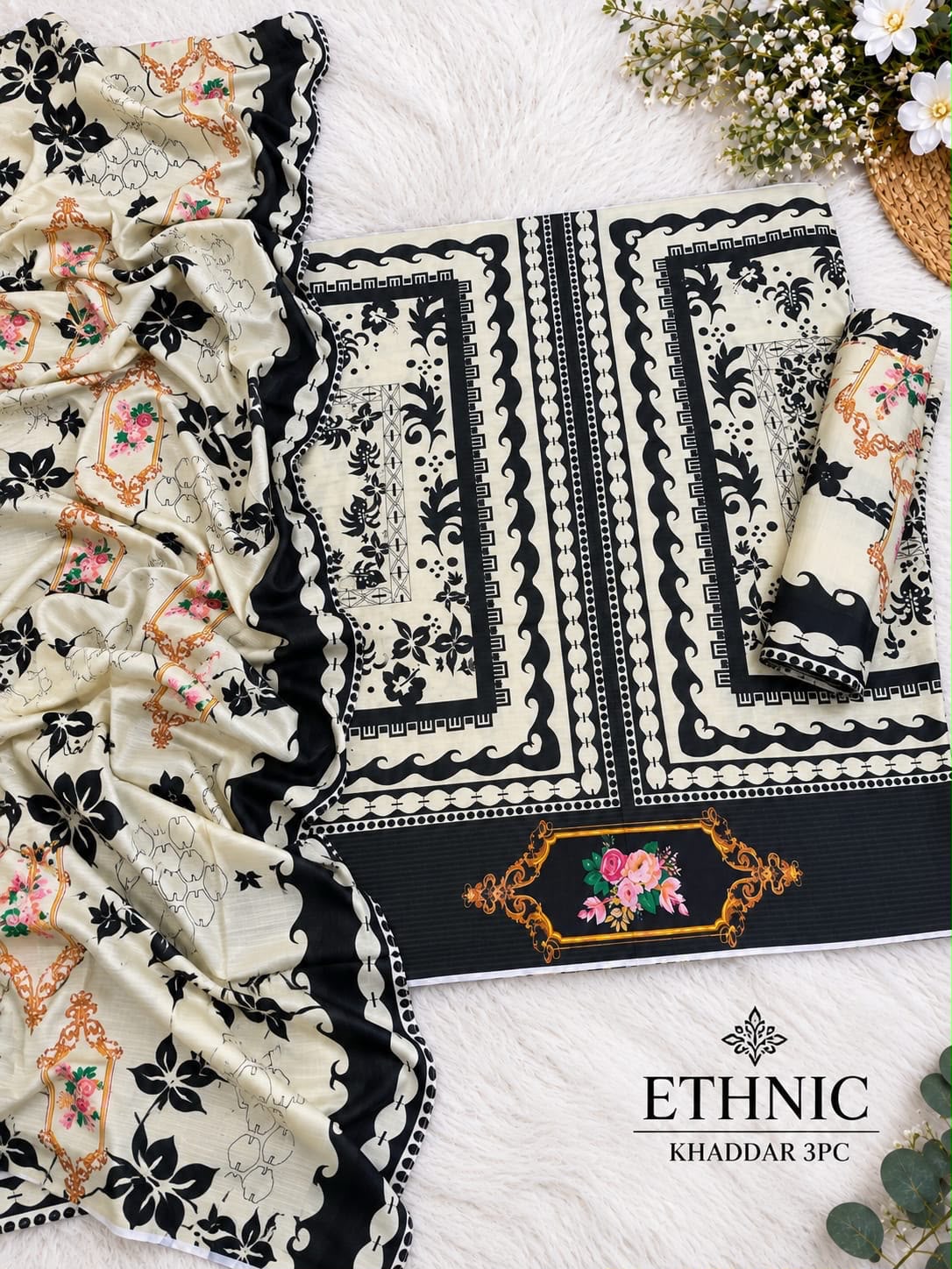 A flat-lay presentation of a 3-piece ethnic Khaddar suit material set, featuring a cream-colored base with black floral and ornamental patterns, a matching dupatta, and a folded bottom piece with a floral embroidered panel. The set is displayed on a white faux fur background with decorative flowers in the corner and the brand text 'ETHNIC KHADDAR 3PC' at the bottom right.