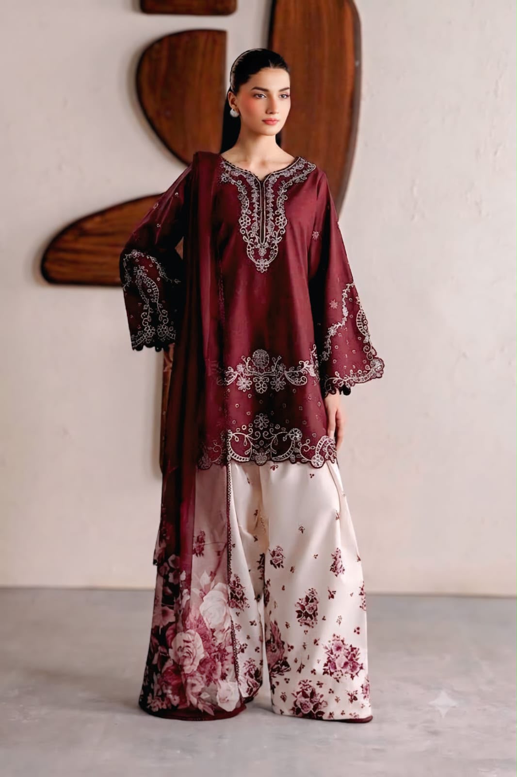 A woman models a traditional South Asian outfit featuring a deep maroon embroidered kurta with wide sleeves and a matching dupatta, paired with wide-leg palazzo pants in cream with a burgundy floral print.