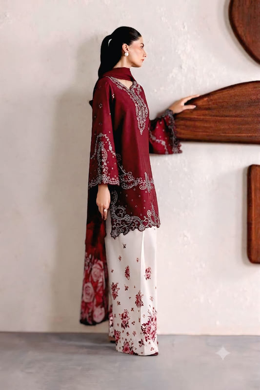 A woman models a traditional South Asian outfit consisting of a deep maroon embroidered kurta with wide sleeves and a matching dupatta, paired with white wide-leg trousers featuring red floral prints. She is standing in profile against a textured off-white wall with wooden wall art.