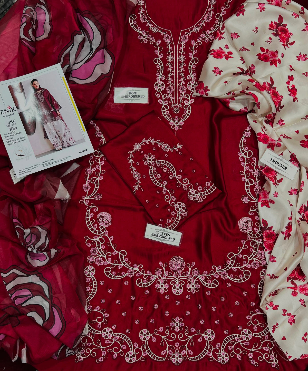 A flat-lay presentation of a women's ethnic wear set, featuring a deep red kurta with intricate white embroidery, a matching red dupatta, a cream-colored fabric with red floral print, and a printed fashion magazine.