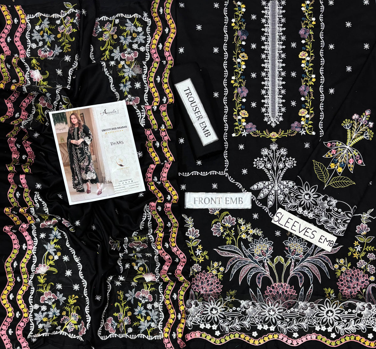 Black embroidered dress fabric laid flat, featuring intricate floral and vine embroidery in white, pink, yellow, and purple. A printed pattern booklet and two white fabric labels reading 'CUFF SKIN CAR', 'FRONT EMB', and 'Sleeves Emb' are placed on top of the fabric.