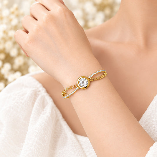 A gold bangle bracelet with a central round clear crystal and delicate chain details, displayed on a soft beige fabric background with dried floral accents.