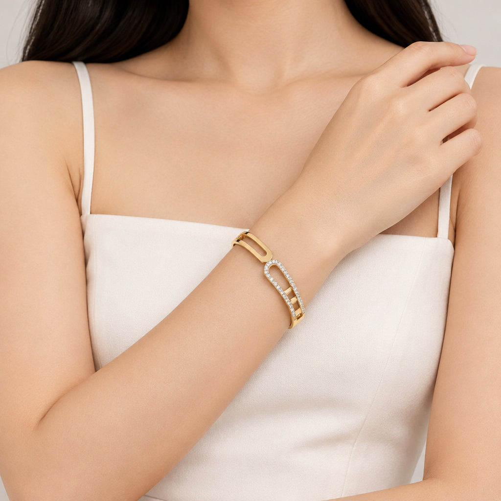 A gold-toned bangle bracelet with a curved openwork design, featuring one side set with small clear stones, displayed on a black surface with cream fabric in the background.