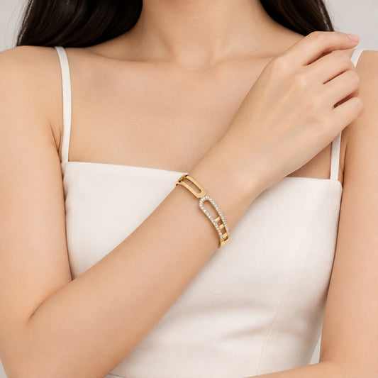 A gold-toned bangle bracelet with a curved openwork design, featuring one side set with small clear stones, displayed on a black surface with cream fabric in the background.