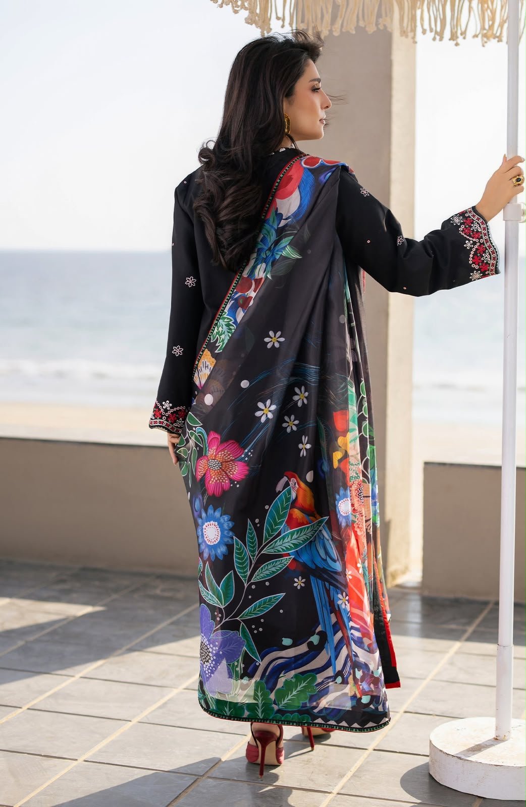 Woman wearing a colorful floral abaya with a black background, standing on a rooftop with ocean view.