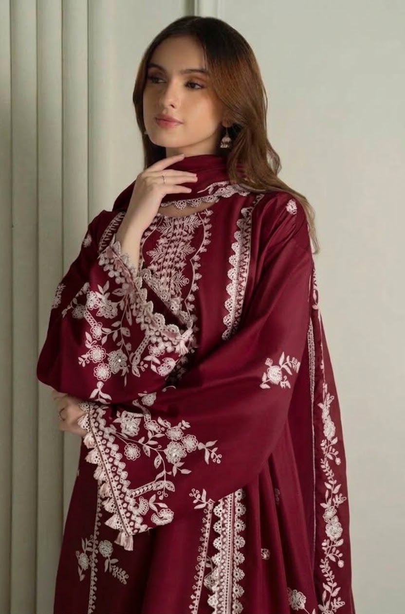 A woman wearing a deep maroon traditional outfit with intricate white floral embroidery, posed against a neutral light gray background.