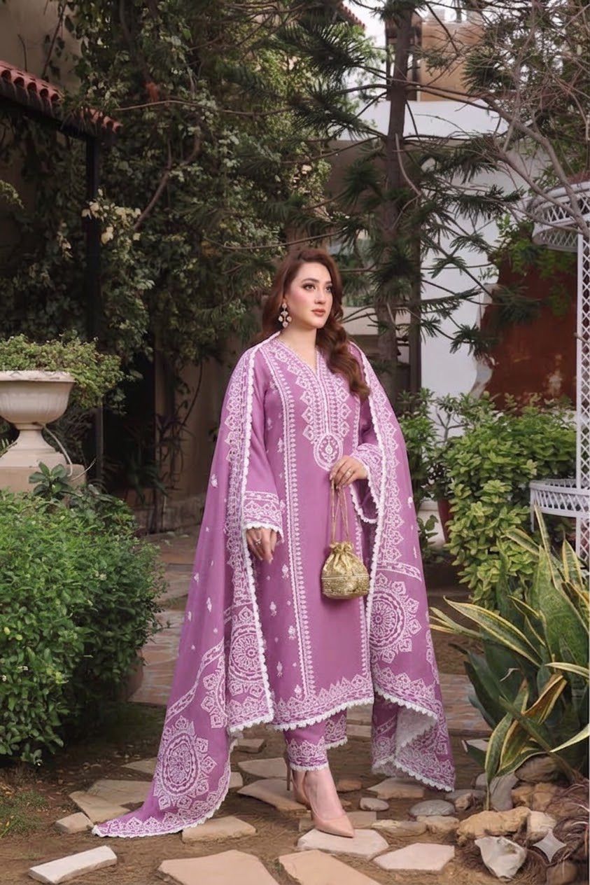 A woman stands outdoors in a garden courtyard wearing a lavender-purple traditional outfit with white floral embroidery and a matching long draped outer layer. She is holding a small gold handbag.