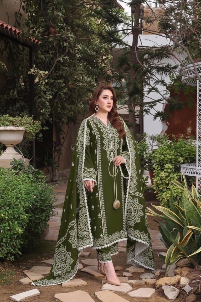 A woman models an olive green ethnic three-piece suit with intricate white embroidery in a garden setting. The outfit includes a long kurta, matching trousers, and a draped dupatta.