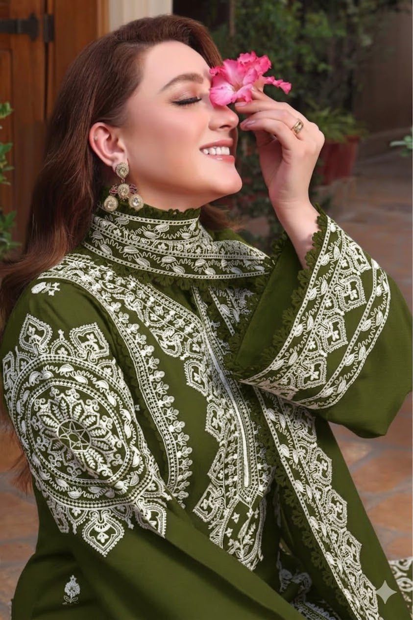 A woman wearing an olive green embroidered traditional outfit with intricate white patterns, holding a pink flower near her face in an outdoor setting.