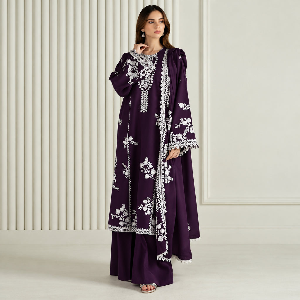 A woman models a deep purple traditional outfit with intricate white floral embroidery and silver mirror work, set against a neutral cream-colored paneled background.
