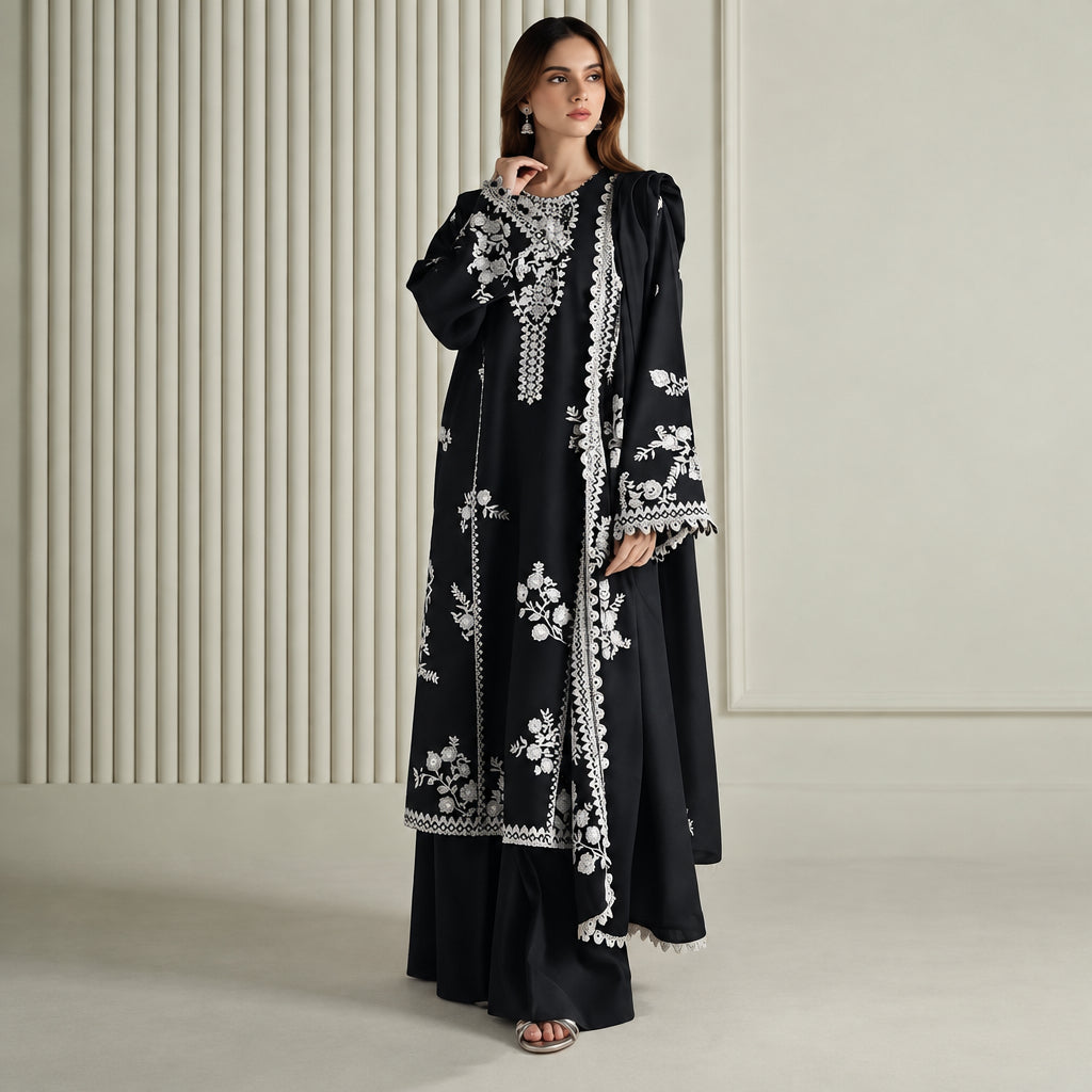A woman modeling a black long kurta with white floral embroidery and white border trim, paired with matching black wide-leg pants, set against a light beige paneled background.