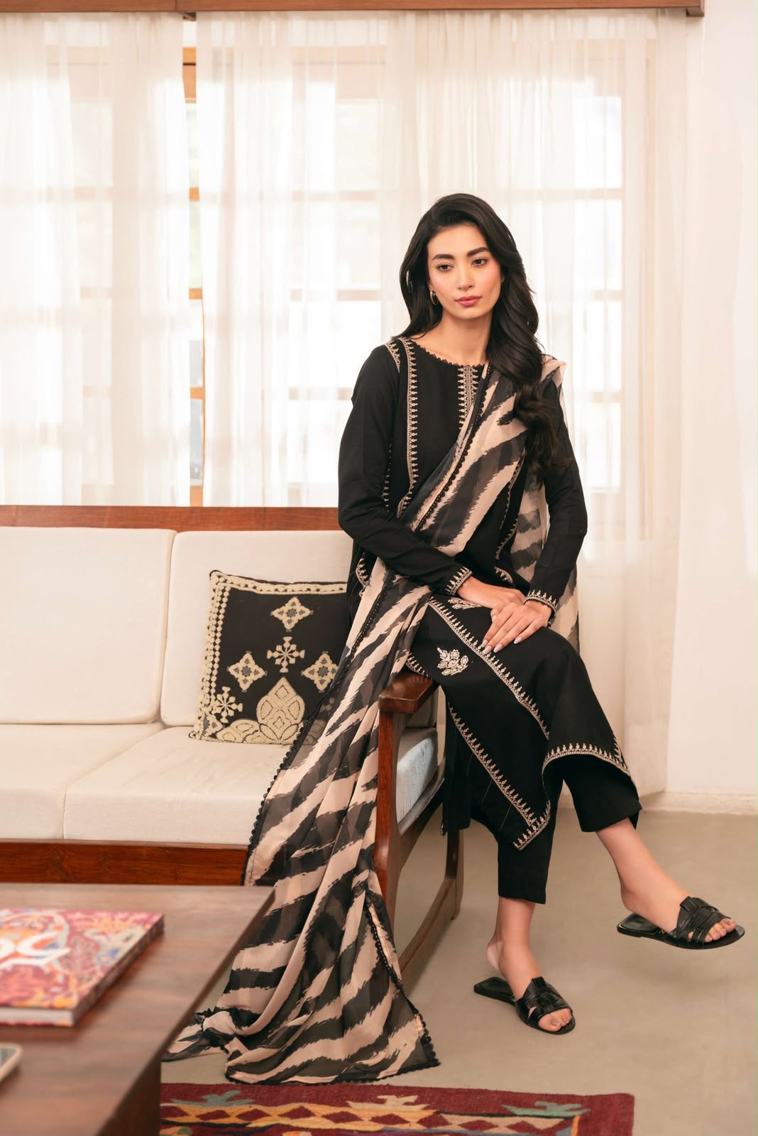 Woman sitting on a couch with a patterned blanket, in a room with white curtains.