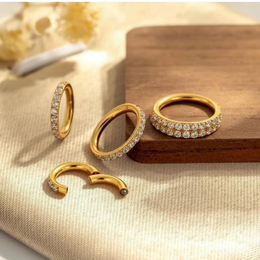 Four gold hoop earrings with clear stones, including two full hoops, one half-hoop, and one open-ended piece, arranged on a beige fabric surface with a wooden block.