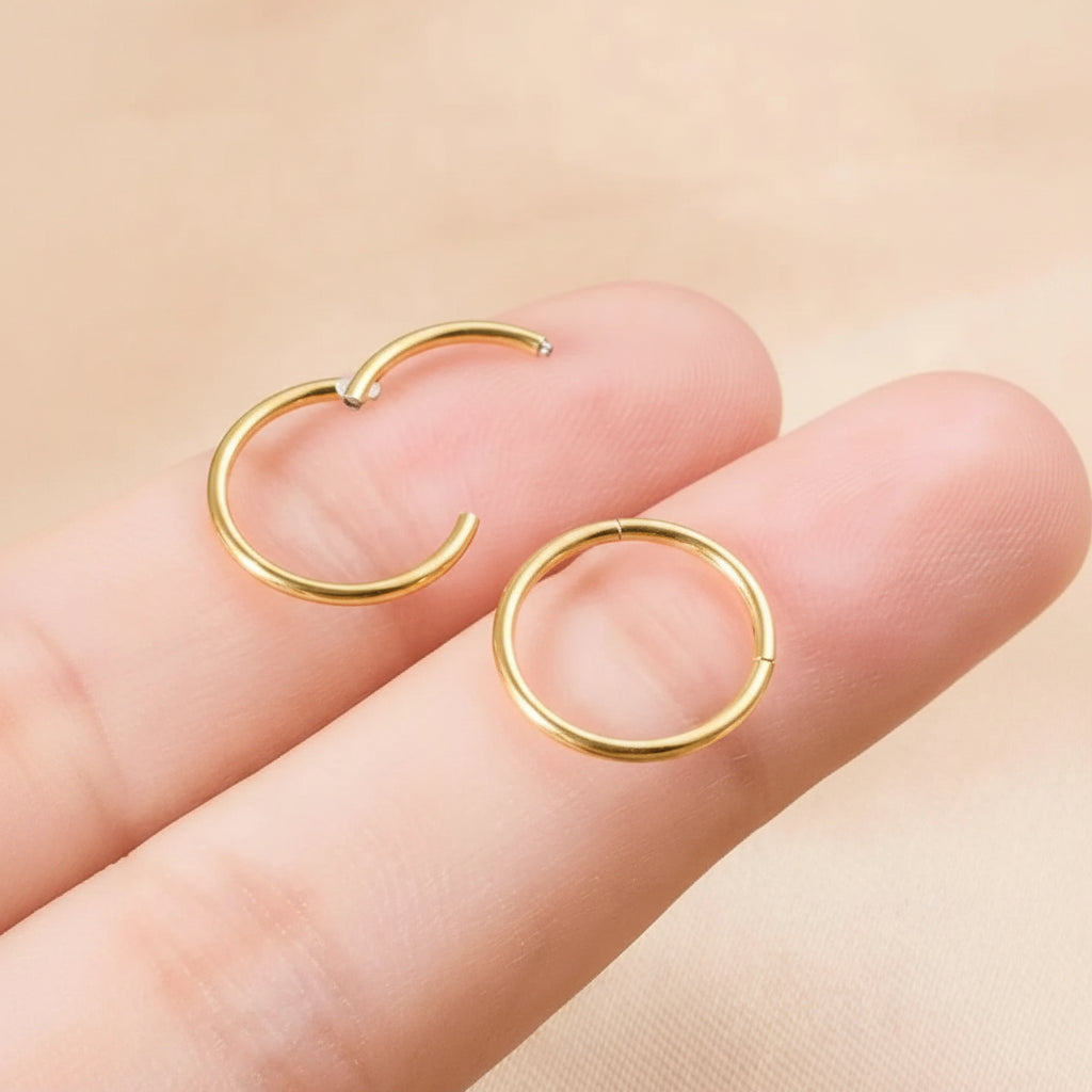 Two small gold hoop earrings held on a person's fingers against a soft beige background.