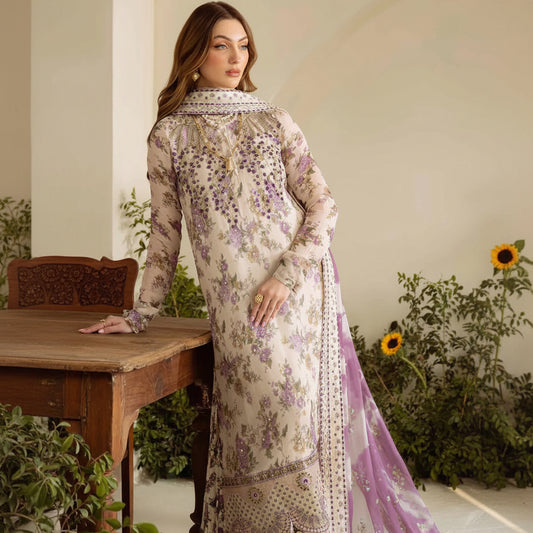 A woman models a traditional long-sleeve ethnic dress with floral embroidery and a matching sheer dupatta, posed beside a wooden table in an indoor setting with plants.