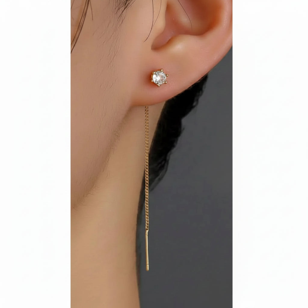 A close-up of a model's ear wearing a delicate gold dangle earring with a clear heart-shaped crystal stud and a long hanging chain.