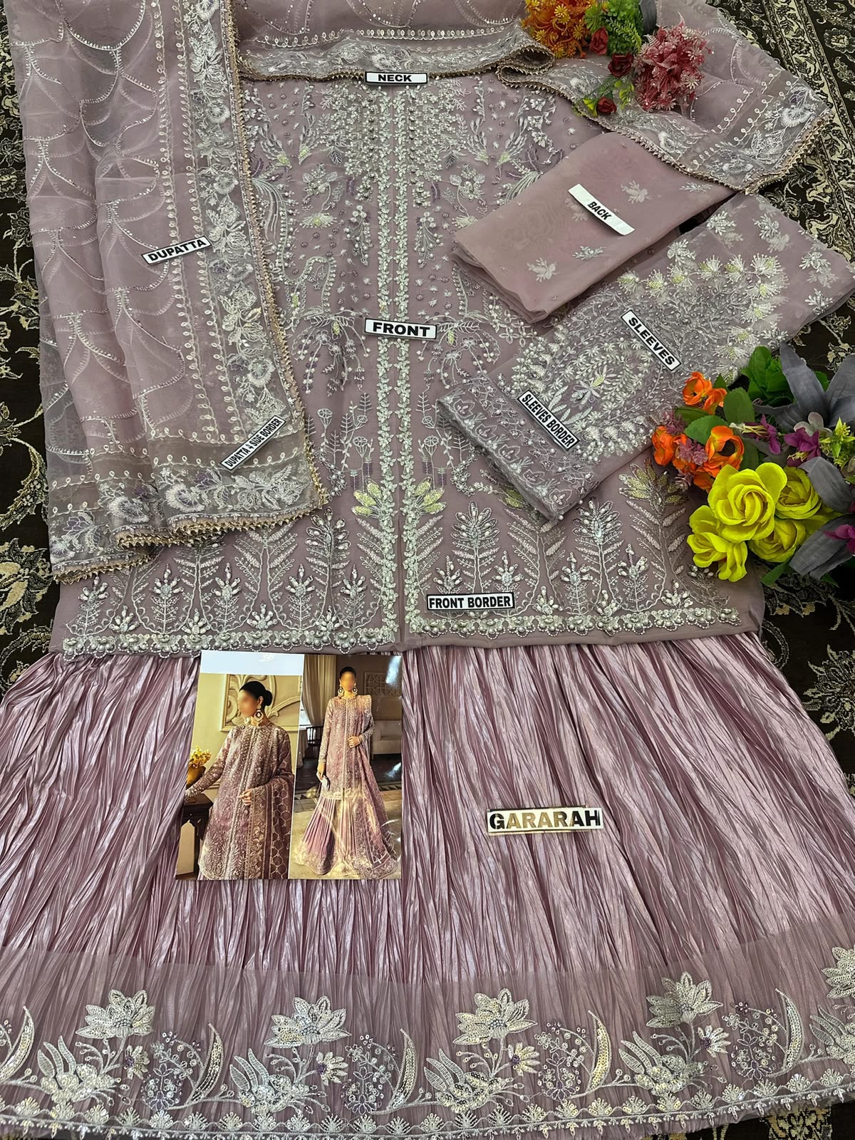 A three-piece women's ethnic suit set in dusty rose pink with intricate silver and white embroidery, featuring a kurta, matching pants, and dupatta, styled with flowers on a dark patterned rug.