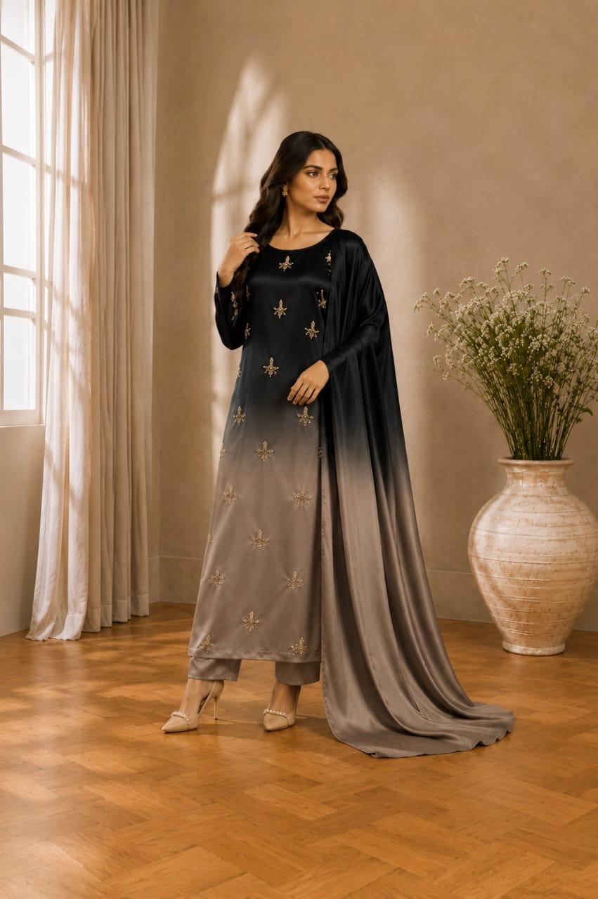 A woman models a traditional Indian outfit consisting of a black and taupe ombre long kurta with gold embroidery, matching trousers, and a draped dupatta, set in a warm, sunlit room with wooden floors and a large vase of flowers.