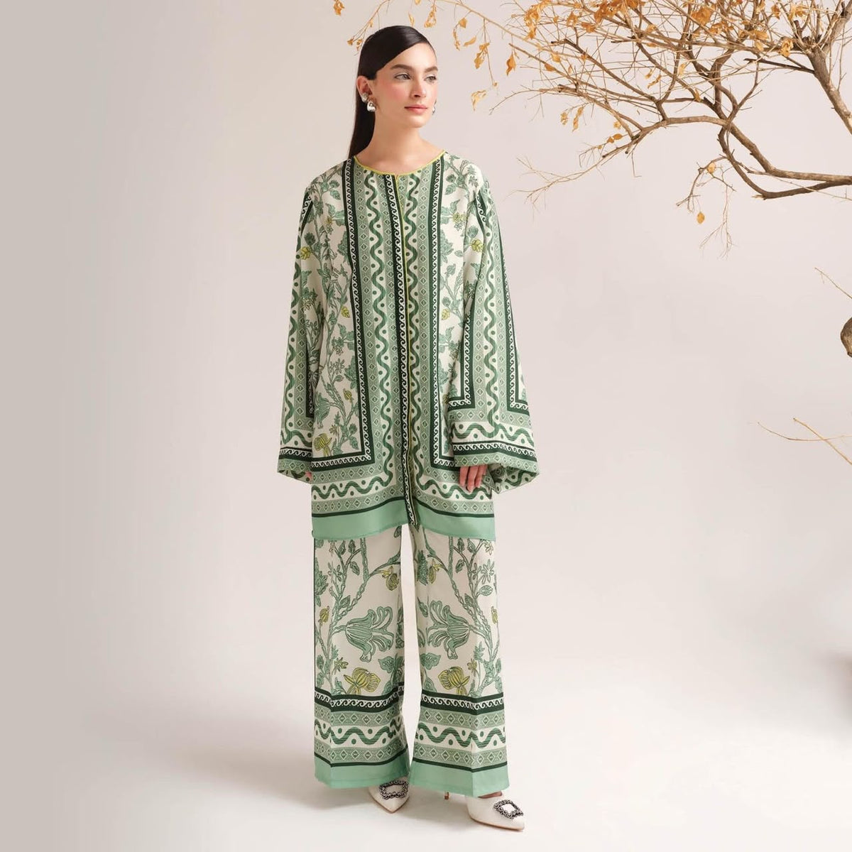 A woman models a light green and white printed two-piece outfit consisting of a long-sleeve kurta and matching wide-leg trousers, styled with white pointed shoes against a neutral studio backdrop with a decorative tree branch.