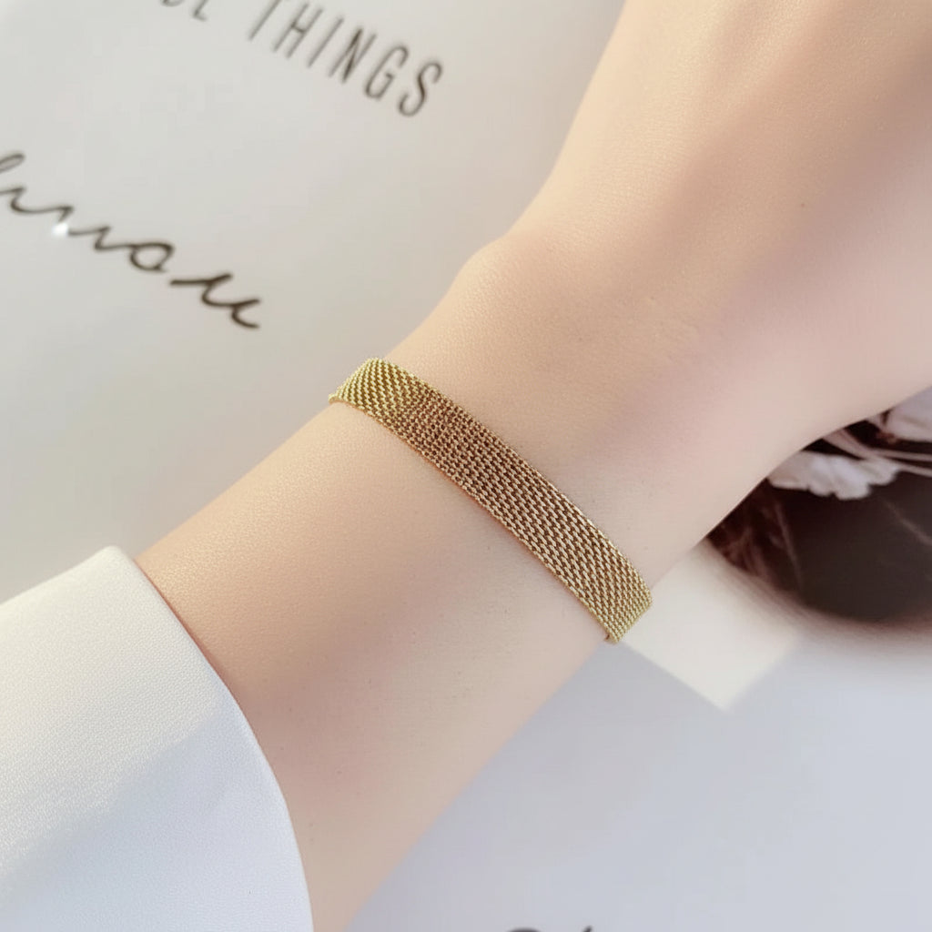A close-up of a delicate gold mesh bracelet worn on a person's wrist, set against a soft white background with partial text.