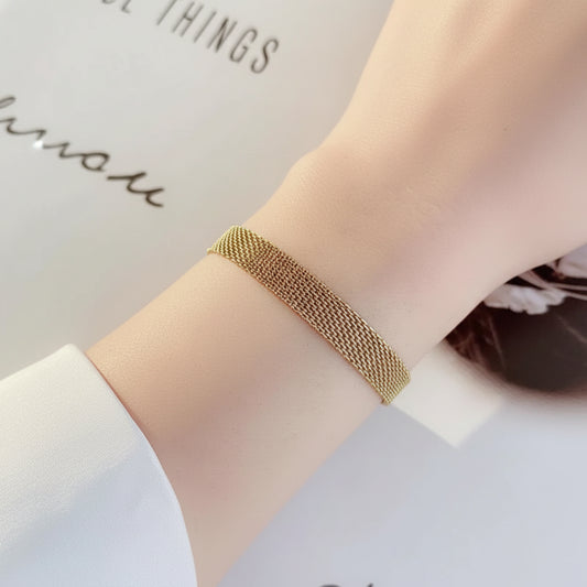 A close-up of a delicate gold mesh bracelet worn on a person's wrist, set against a soft white background with partial text.