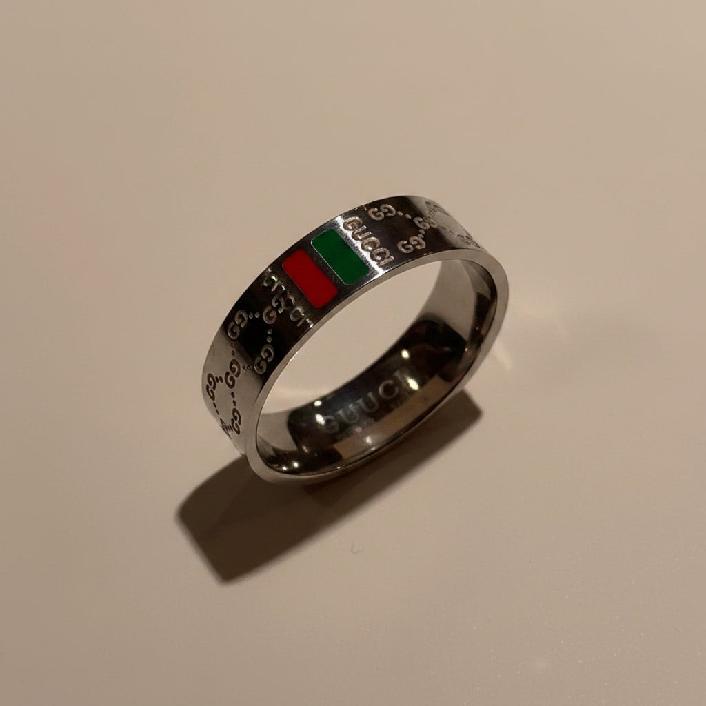 A silver-toned Gucci ring featuring the brand's signature GG monogram pattern and a green and red accent stripe, displayed on a beige surface.