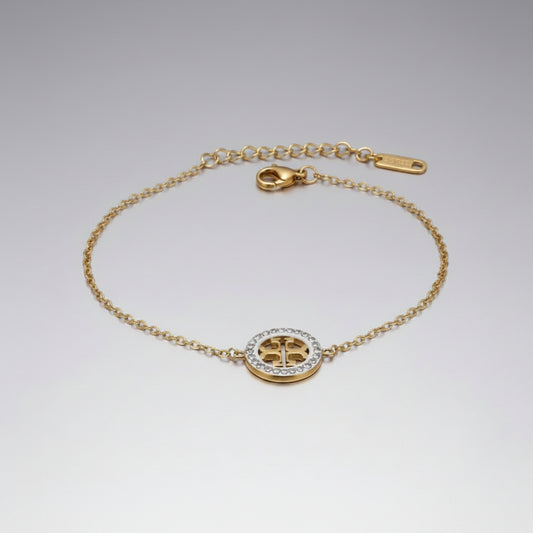 A gold chain bracelet with a circular logo charm, featuring a gold Tory Burch-style monogram center surrounded by a halo of small clear stones, displayed on a white fabric background.