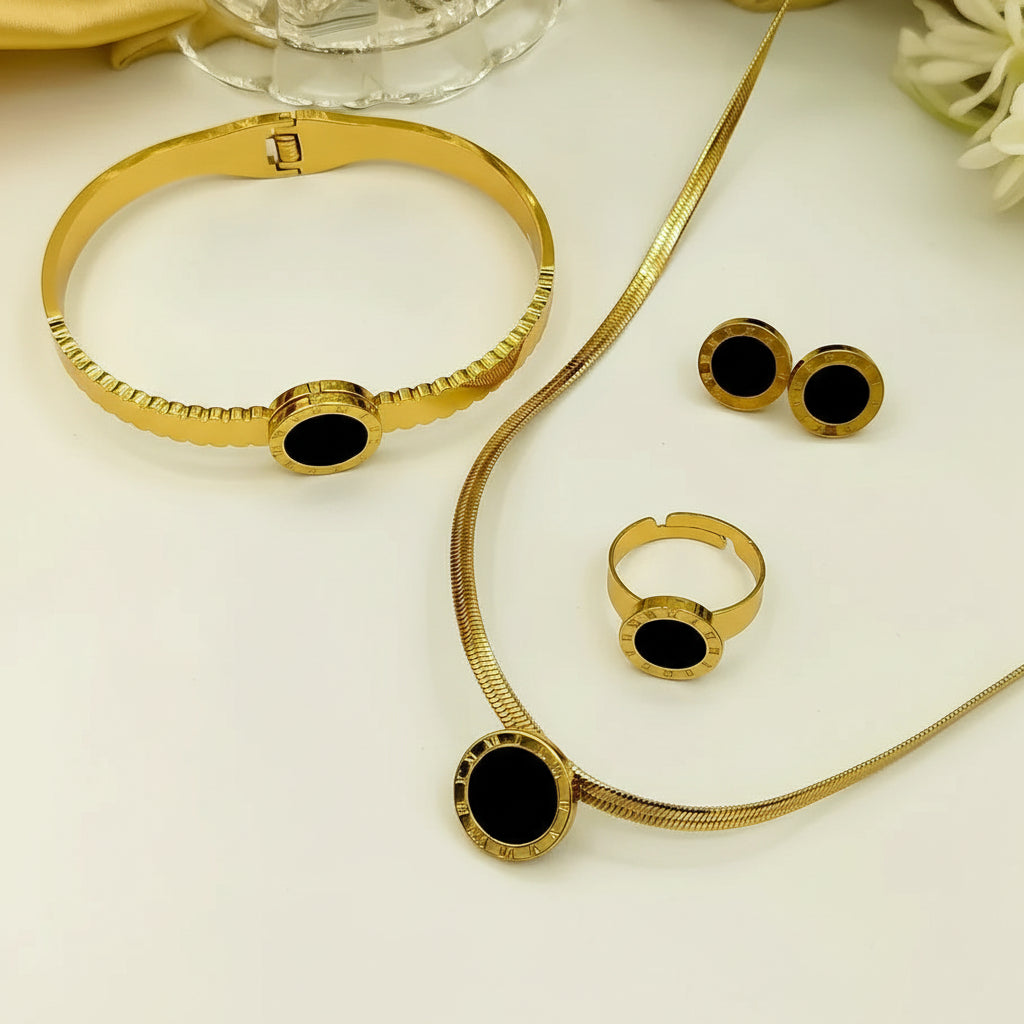 Gold jewelry set with black oval inlays, including a bracelet, necklace, ring, and earrings, arranged on a white surface with a glass bowl and yellow fabric in the background.