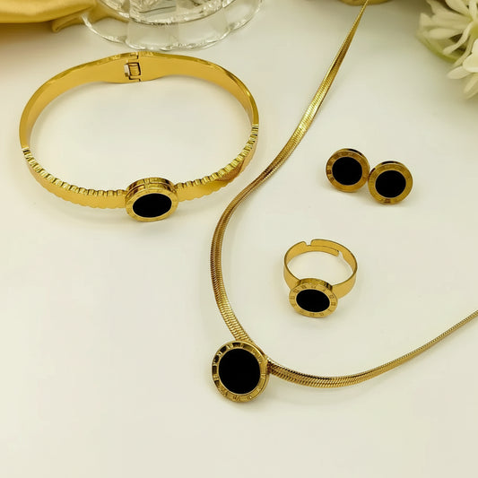 Gold jewelry set with black oval inlays, including a bracelet, necklace, ring, and earrings, arranged on a white surface with a glass bowl and yellow fabric in the background.