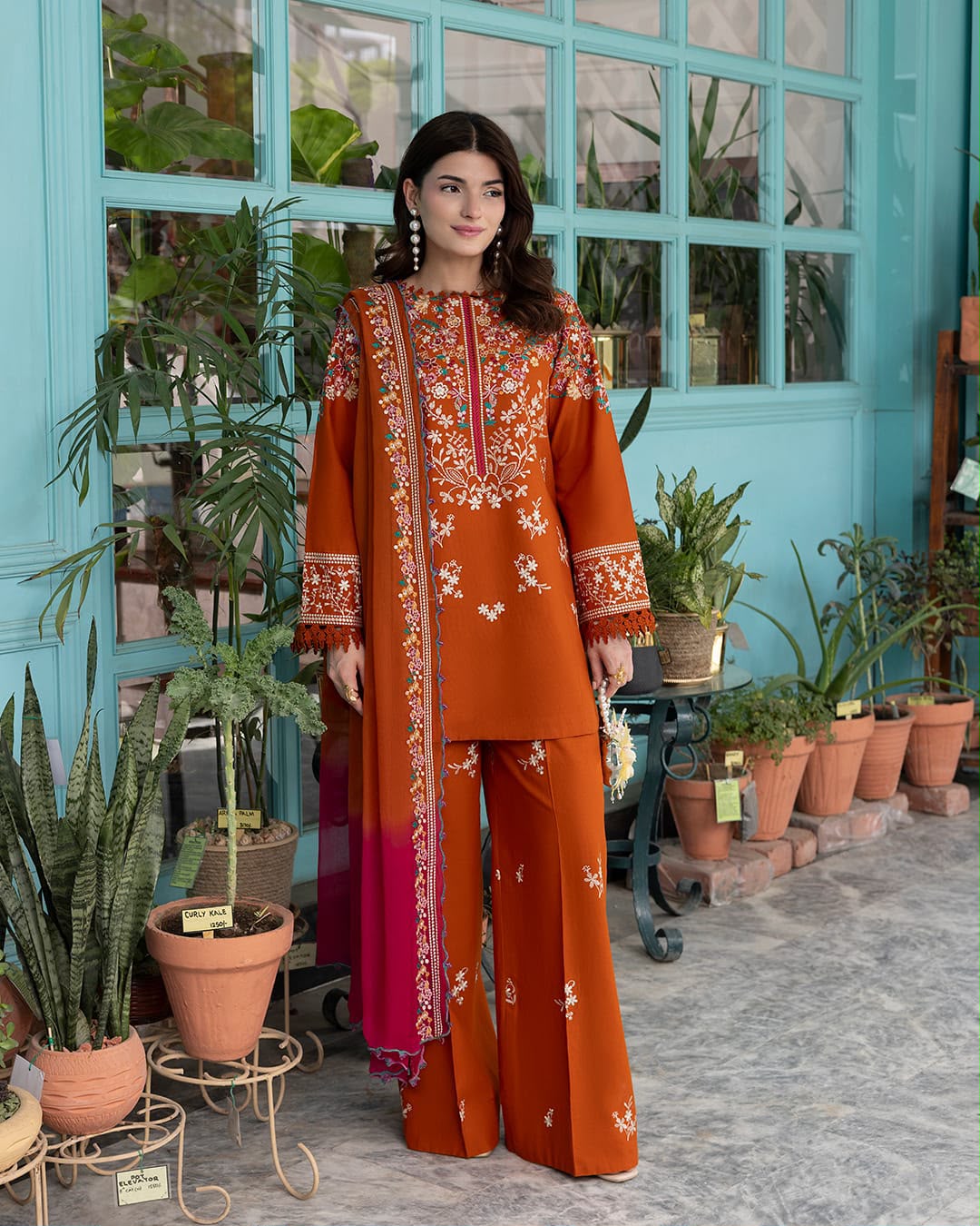 A woman models a three-piece orange kurta set with intricate floral embroidery and a magenta gradient dupatta, set against a turquoise windowed wall with potted plants.