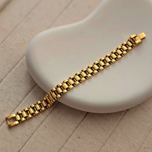 A gold-tone metal link bracelet with a polished finish, laid diagonally across a light beige textured surface and a white curved display prop.