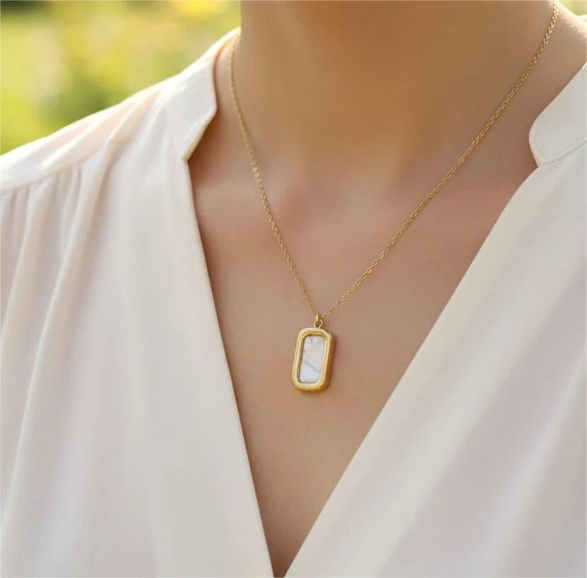 A gold rectangular pendant necklace with a white inlay, worn by a model in a white blouse against a softly blurred green background.