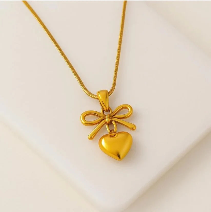 A gold necklace with a bow and heart pendant, displayed on a clean, light beige surface.