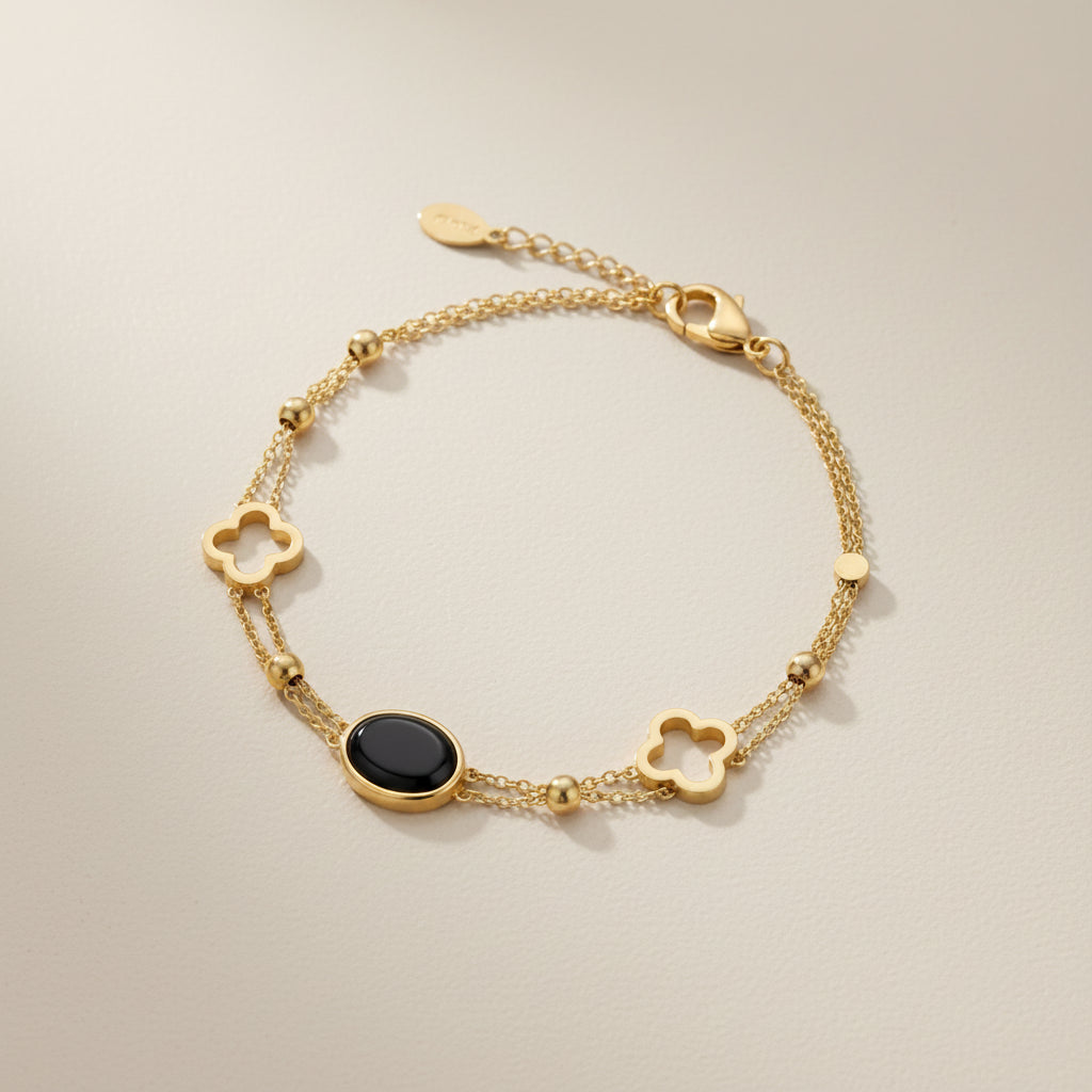A gold chain bracelet featuring three clover-shaped charms, a small round gold bead, and a central oval black onyx stone, displayed on a textured gray surface.