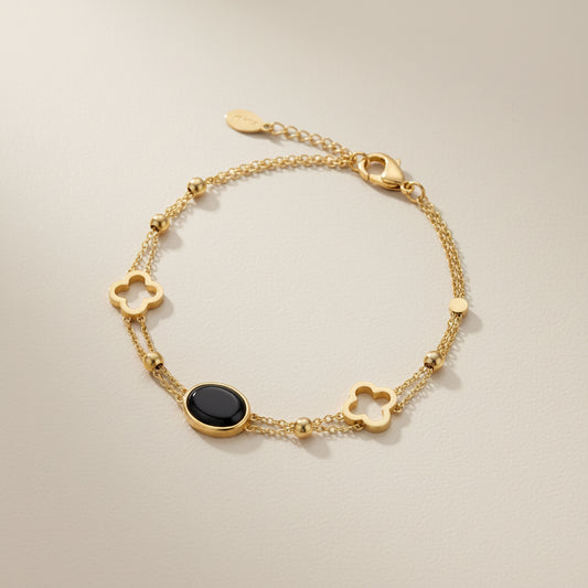 A gold chain bracelet featuring three clover-shaped charms, a small round gold bead, and a central oval black onyx stone, displayed on a textured gray surface.
